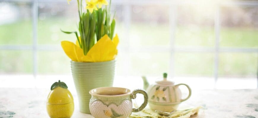 A table in front of a large window set with a tea pot, tea cup, ceramic lemon, and a vase with flowers